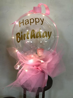 Happy birthday print on the transparent balloon white net wrapping and gold bow in a box with string lights