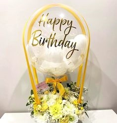Transparent Balloon Printed Happy Birthday in gold stuffed with 4 white balloons Tied with ribbons to a basket of 12 yellow and white flowers